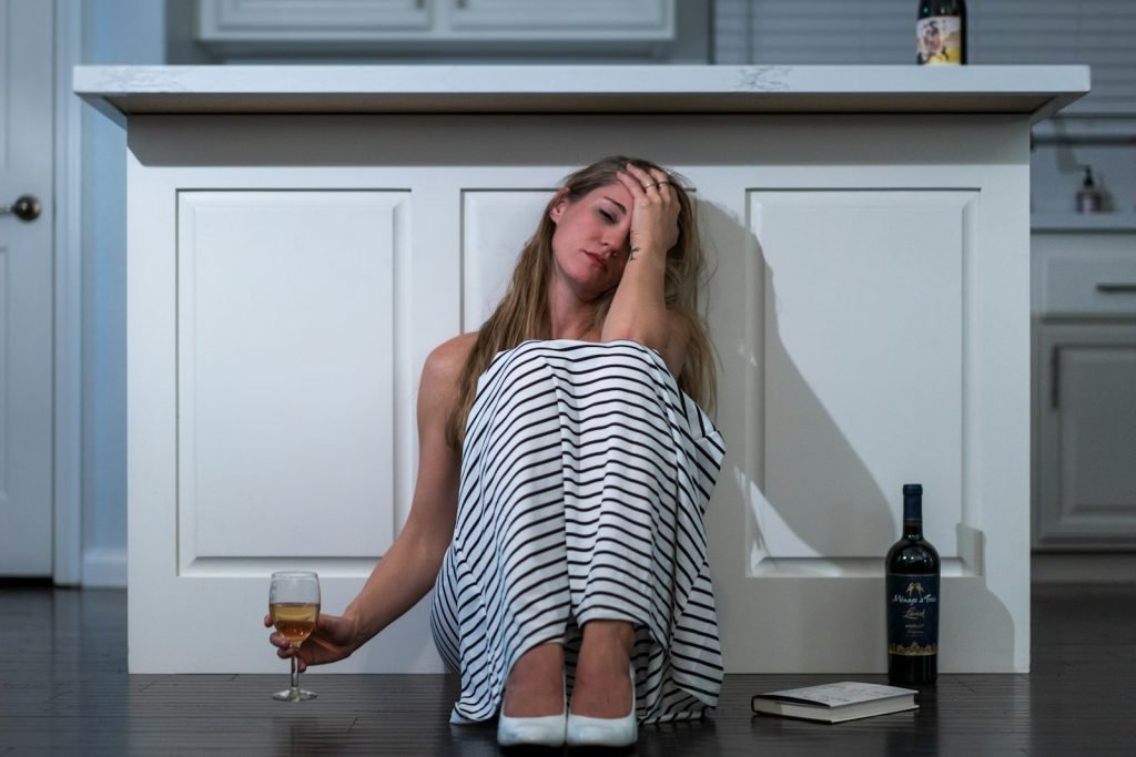 Pessoa sentada no chão da cozinha ao lado de uma taça e uma garrafa de vinho